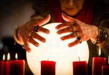 A person with adorned hands hovering over a glowing crystal ball surrounded by candles
