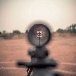 Rifle scope aimed at a distant target outdoors.