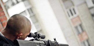 Person aiming a rifle from a building balcony.