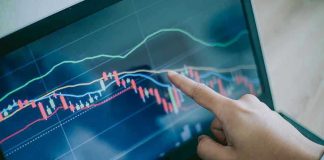 Hand pointing at stock market graph on laptop screen.