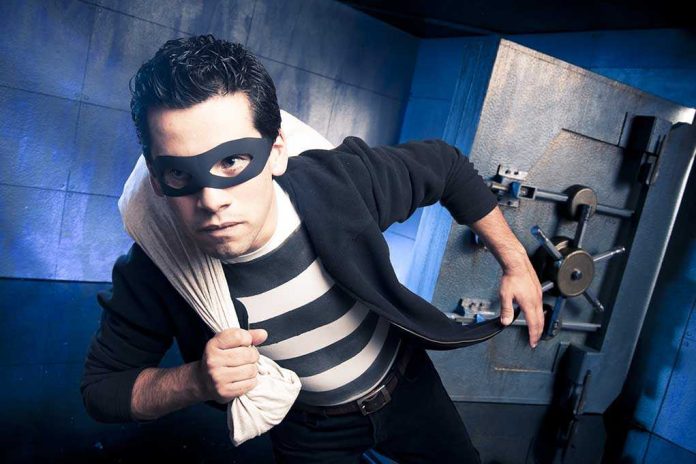 A masked thief in a striped shirt holding a bag while emerging from a bank vault