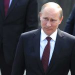 A serious-looking man in a suit walking outdoors with security personnel in the background