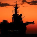 RED ALERT — Gulf Erupts ONCE AGAIN! Silhouette of a naval aircraft carrier against a vibrant sunset