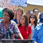 Speaker at outdoor press conference with crowd and protest signs