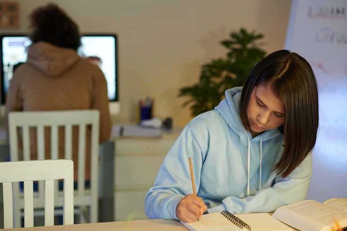 Student in blue hoodie writing in notebook at desk