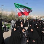 BRUTAL — Iran’s HAIR POLICE Goes High-Tech Group of women in black attire marching with an Iranian flag