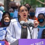Brutal Call-Out — AOC Laughs, Families GRIEVE Woman speaking at outdoor rally behind microphones