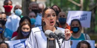 Brutal Call-Out — AOC Laughs, Families GRIEVE Woman speaking at outdoor rally behind microphones