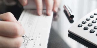 Husband Wrote One Man 70 Checks — The Reason Landed In Federal COURT Person writing a check on a reflective surface with a calculator nearby