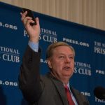Man in suit speaking at podium holding up a smartphone