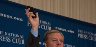 Man in suit speaking at podium holding up a smartphone