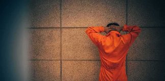 Person in orange jumpsuit stands facing a wall with hands on head