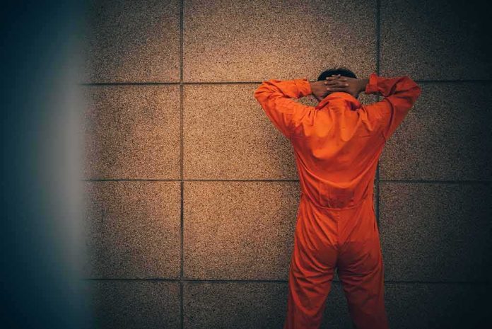 Person in orange jumpsuit stands facing a wall with hands on head