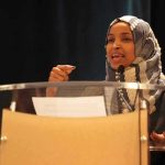Feds At Omar’s Door — Remember What She CLAIMED? Ilhan Omar speaking at a podium with a microphone
