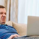 Man grimacing while using a laptop on a couch