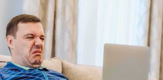 AOC’s 100-Hour Plan Is SICKENING — Look WHO It Covers Man grimacing while using a laptop on a couch