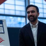 Mamdani’s Staff Coached 200 Influencers — What They Got CAUGHT At Zohran Mamdani smiling indoors near a voting sign with an American flag
