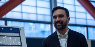 Mamdani’s Staff Coached 200 Influencers — What They Got CAUGHT At Zohran Mamdani smiling indoors near a voting sign with an American flag
